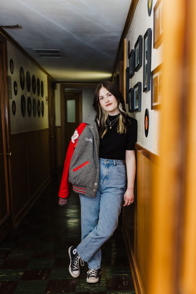 Senior photo of girl in black shirt, letterman jacket, and jeans leaning on record gallery wall in Mauldin Productions recording studio in Gladewater, TX