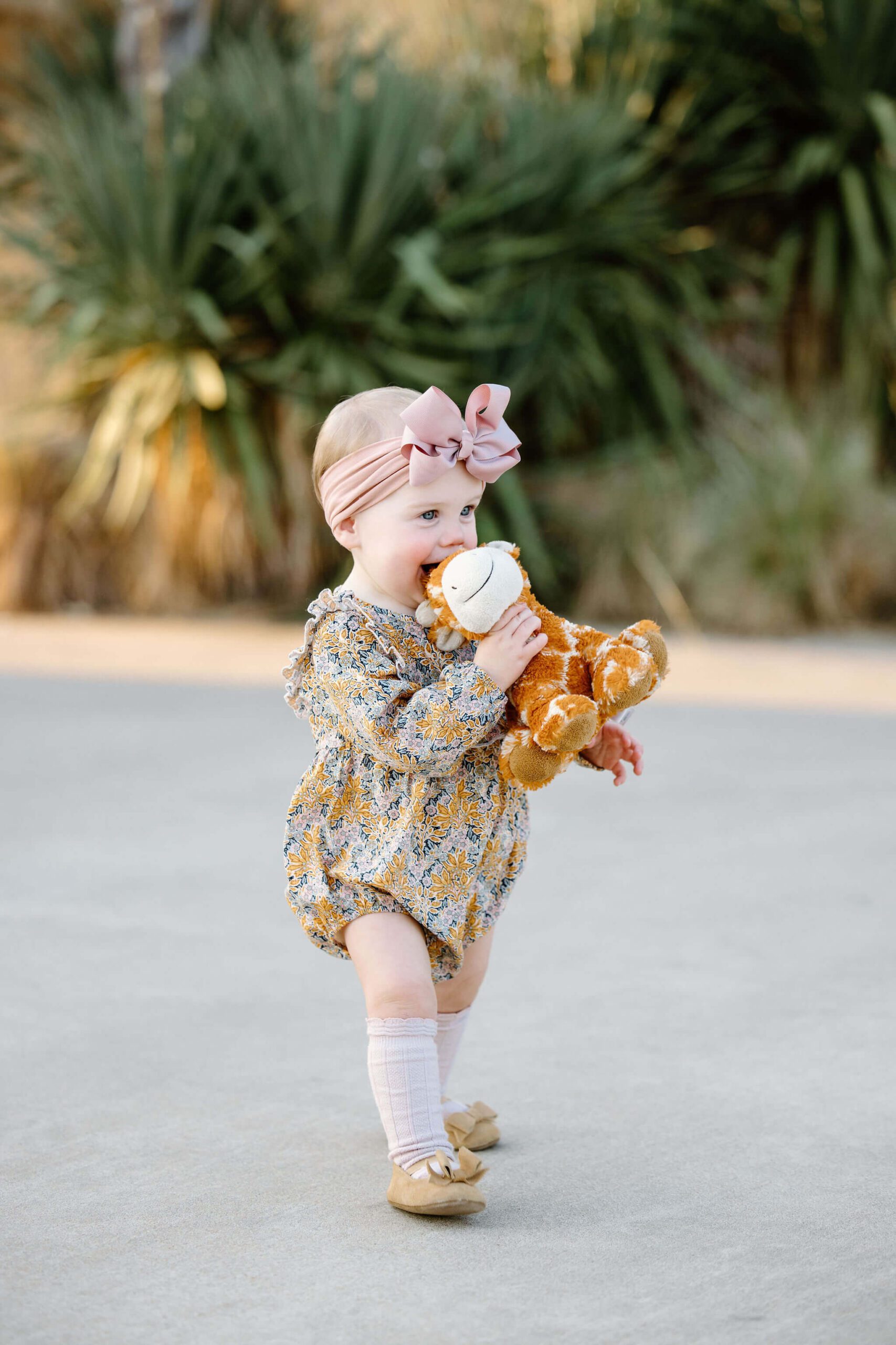 candid baby picture of 1 year old girl in fall colored romper and matching bow holding favorite stuffed animal