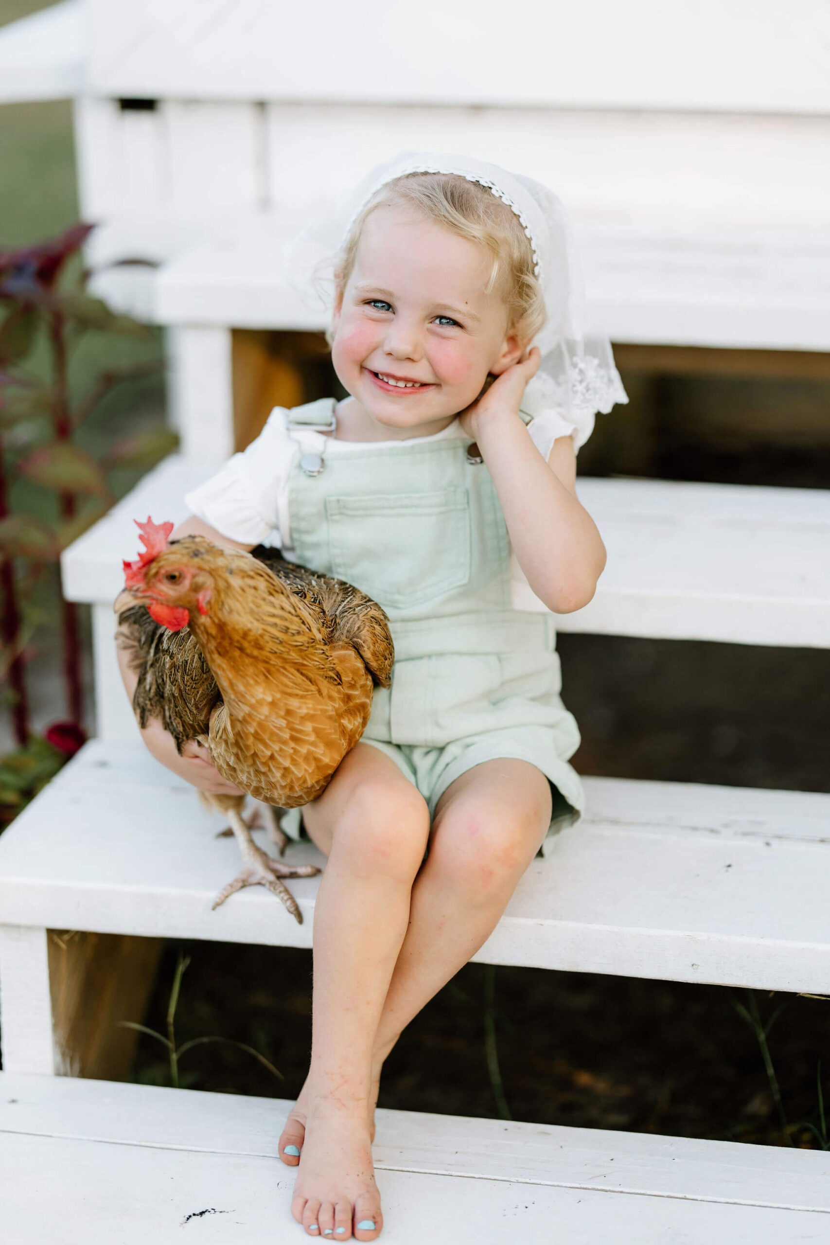 lifestyle photo of toddler girl in green overalls, white shirt, and white bonnet holding chicken on East Texas farm