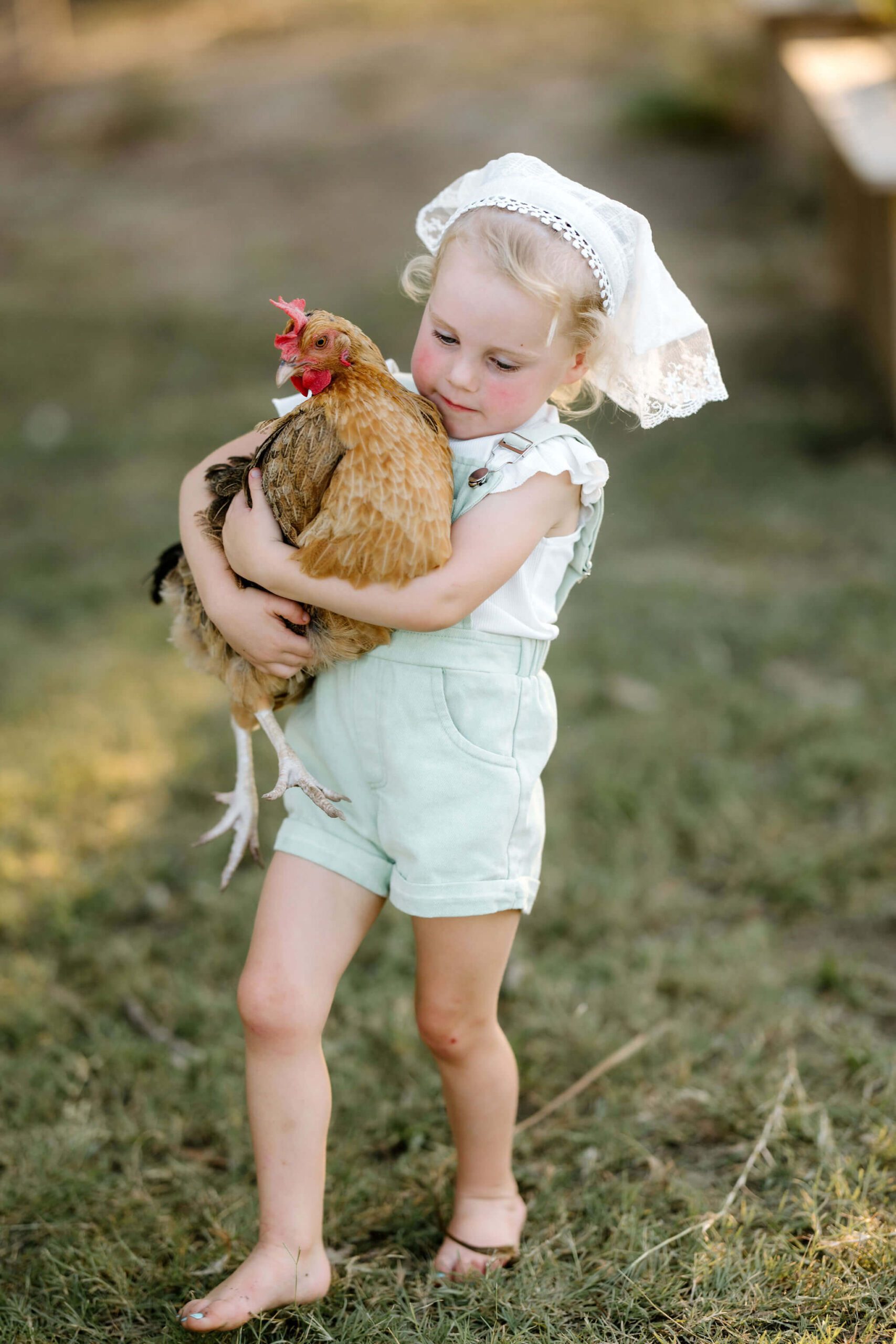 lifestyle photo of toddler girl in green overalls, white shirt, and white bonnet carrying chicken on East Texas farm