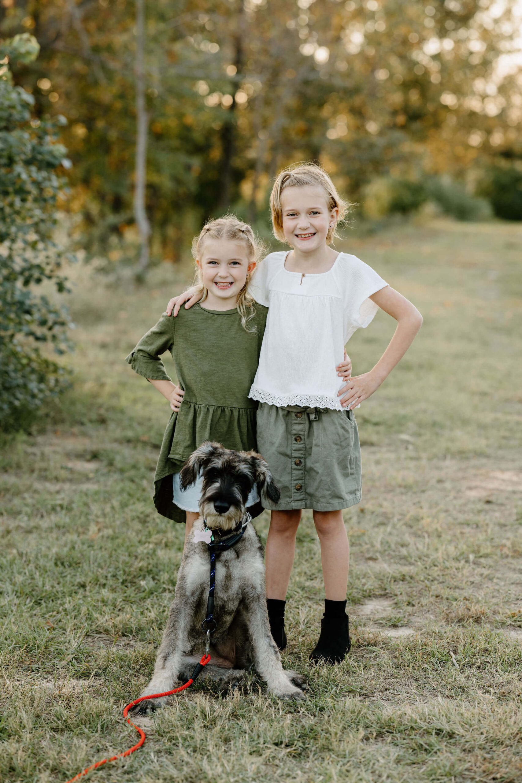 family picture of 2 young sisters and family dog standing in green field