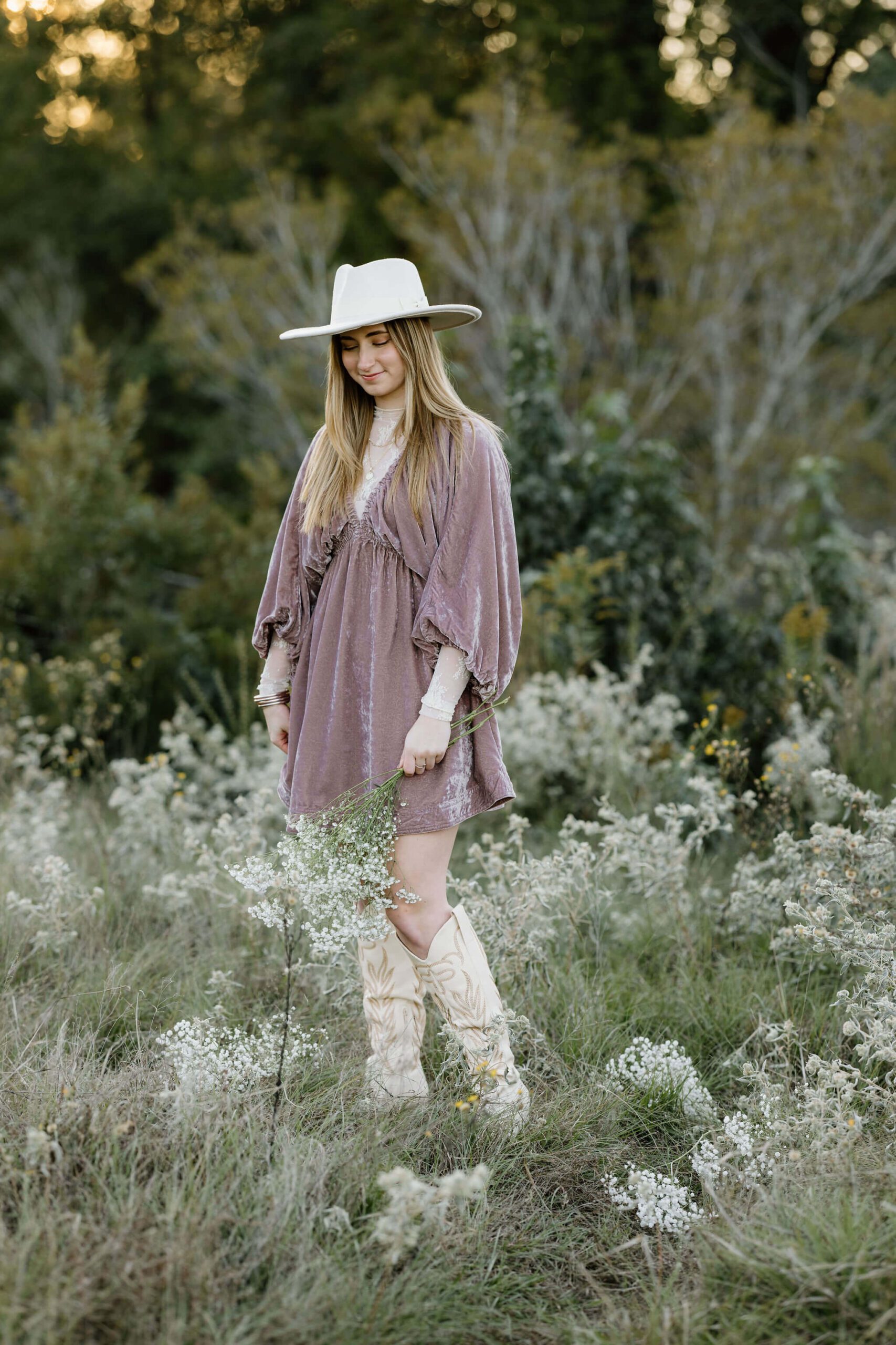 Senior girl in white hat, pink dress, and white boots walking through open East Texas field