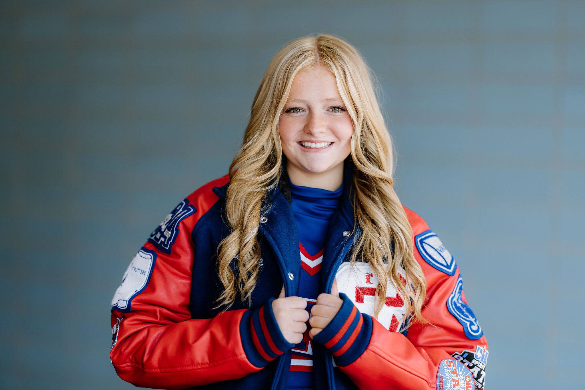 blonde senior girl in high school letterman jacket posing in front of blue wall