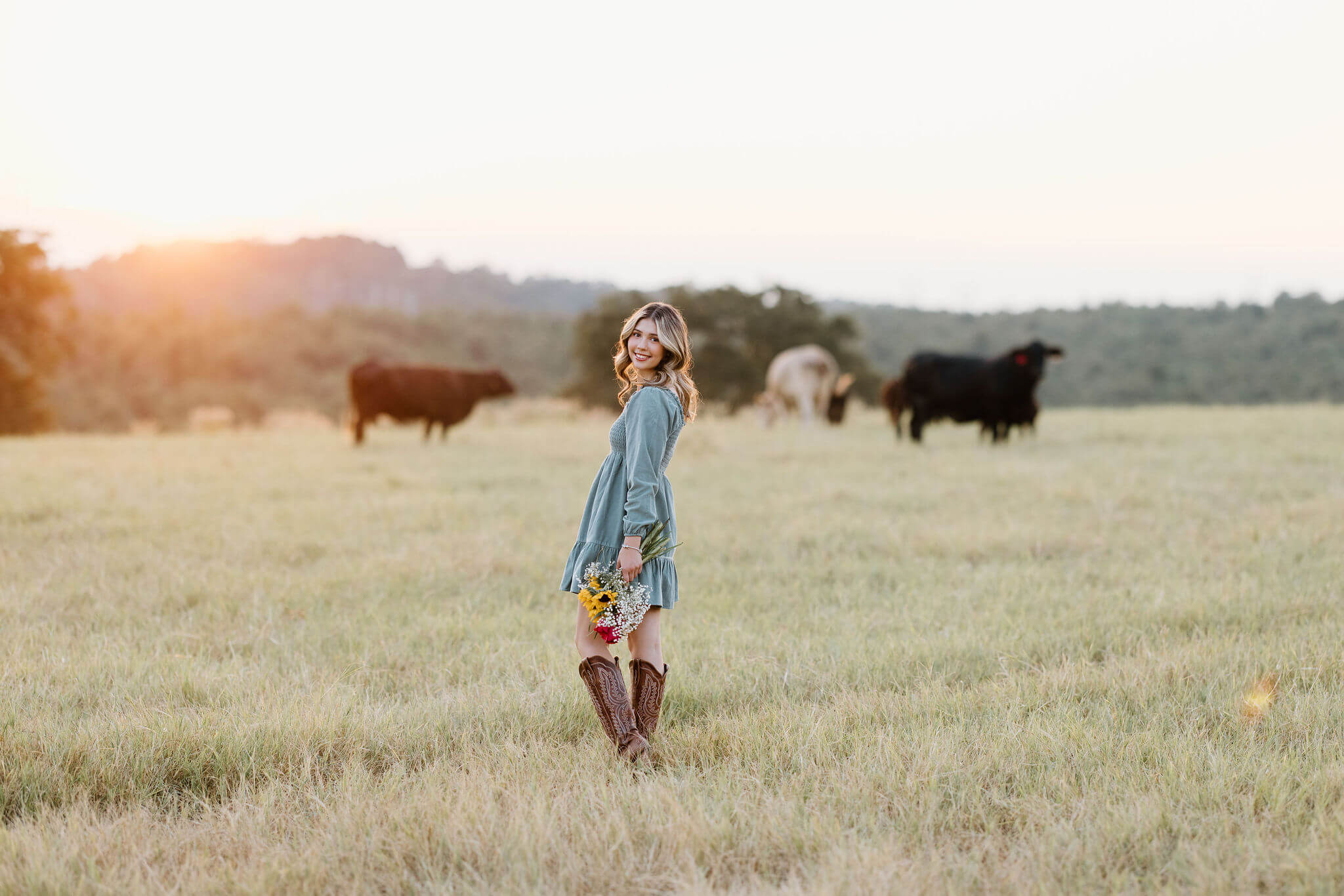 senior picture of girl in teal dress and boots walking through a field at sunset holding a bouquet of flowers