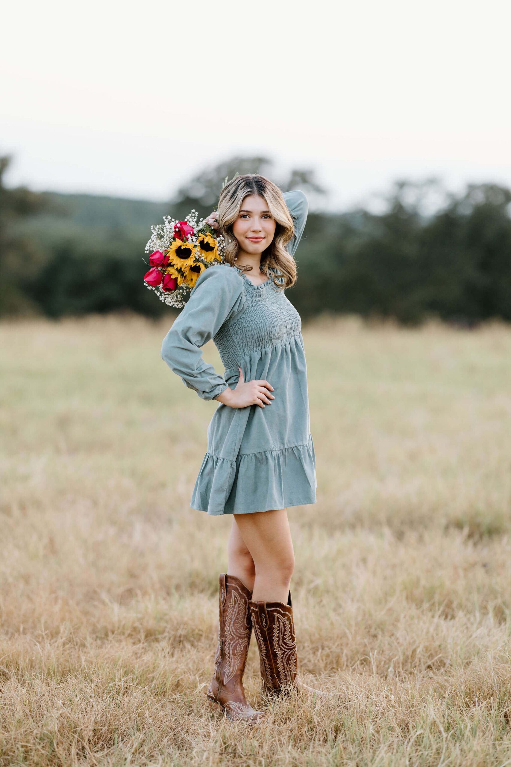 senior girl in dress and cowgirl boots holding a bouquet of colorful flowers in East Texas pasture at sunset