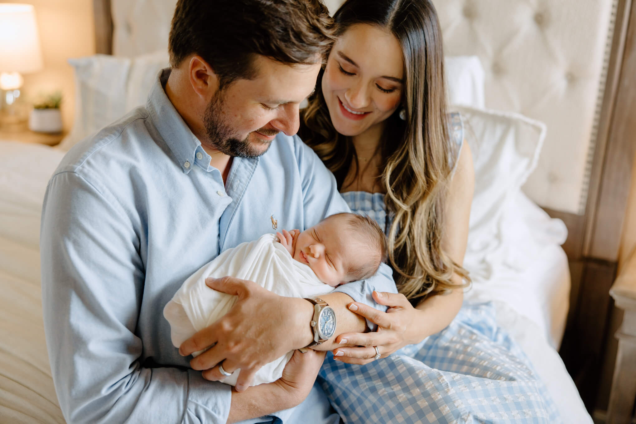 New parents holding newborn son while sitting on the edge of bed in master bedroom